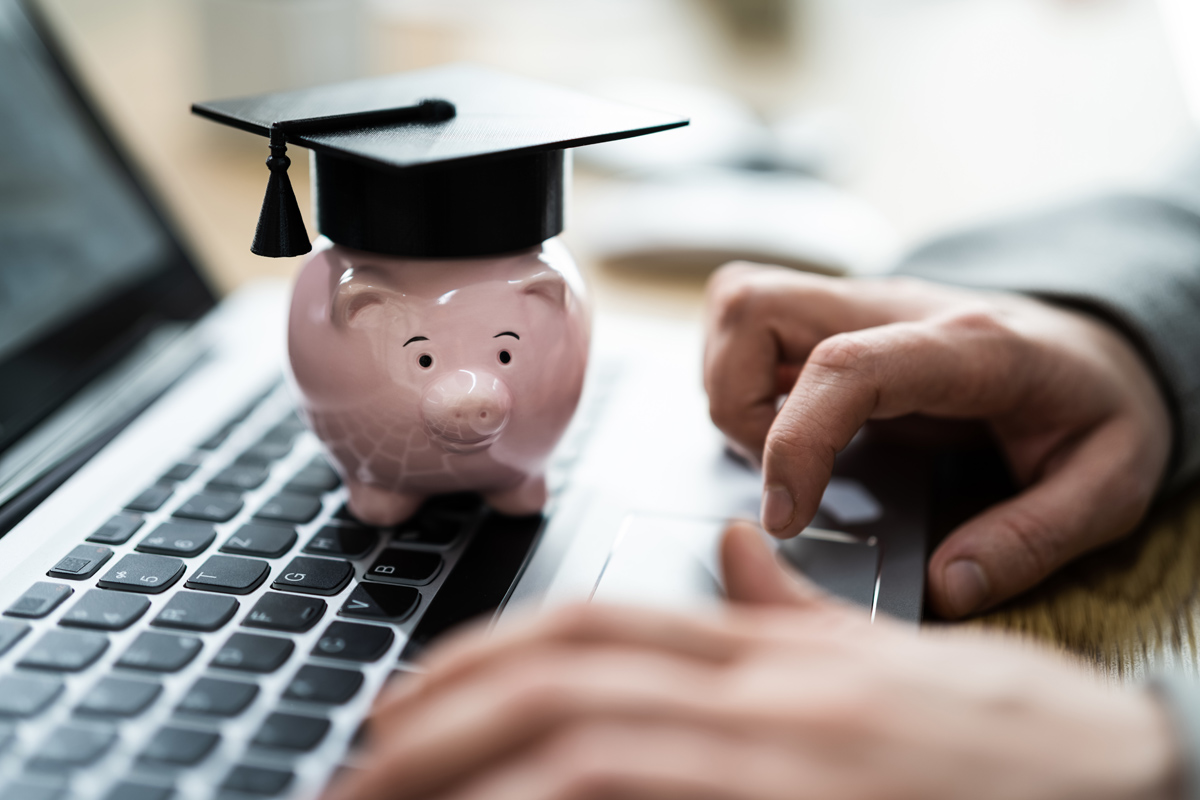 closeup image of hands using a laptop while a small piggybank wearing a graduation cap rests on the keyboard