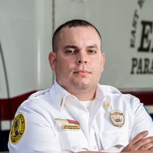 A clean shaven Caucasian man dressed in a white EMS uniform looks stoically into the camera.