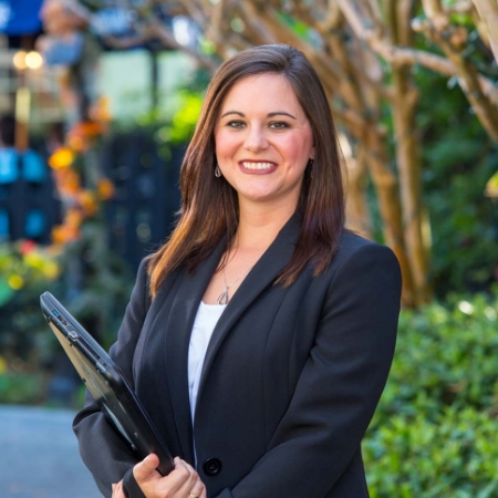 A young white woman dressed in a business suit holding a laptop outside an office building