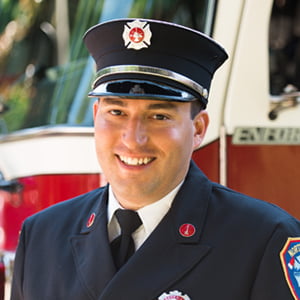 A clean shaven Caucasian man in a fire dress uniform smiles into the camera.