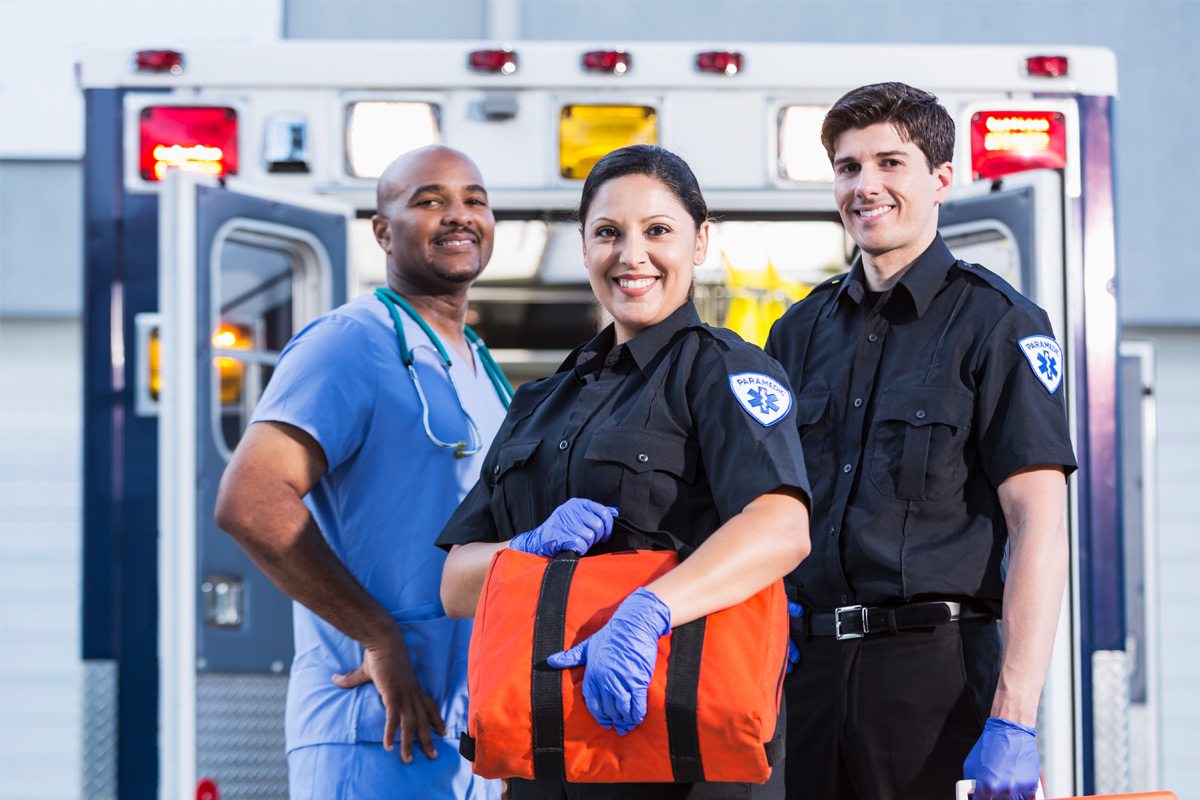 Three first responders smile at the camera as they stand in front of an ambulance